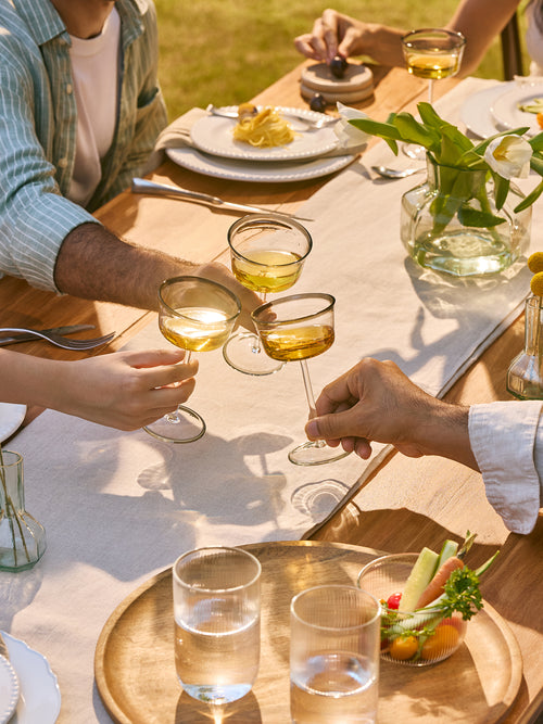 A lifestyle dining scene featuring three people sitting around a wooden table outdoors, clinking coupe glasses filled with white wine. The table is decorated with a white runner, plates, a glass vase with white flowers, and a wooden serving tray holding two glasses of water and a small fruit bowl.