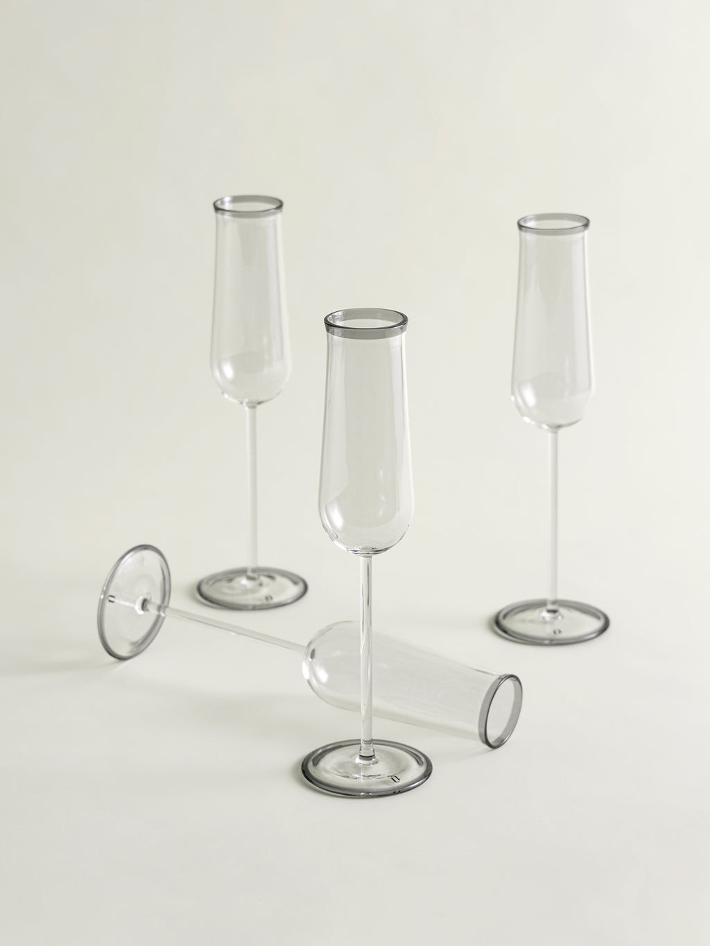 Three clear glass champagne flutes with smoke grey rim and circular bases are arranged on a light neutral studio background, with one flute lying on its side in front.