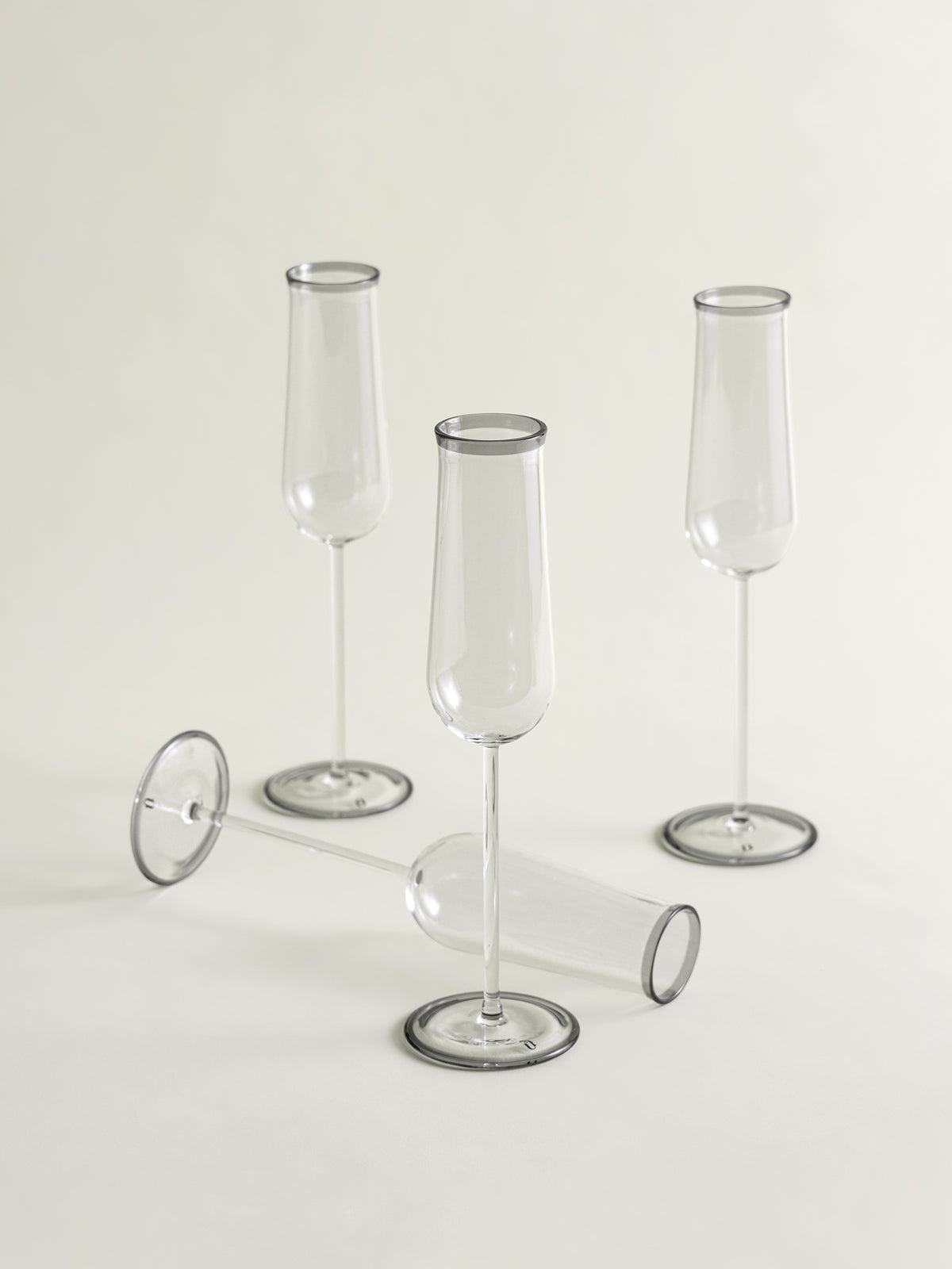 Three clear glass champagne flutes with smoke grey rim and circular bases are arranged on a light neutral studio background, with one flute lying on its side in front.