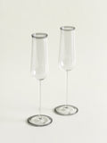 Two clear glass champagne flutes with a subtle gray rim, standing on a light cream background.