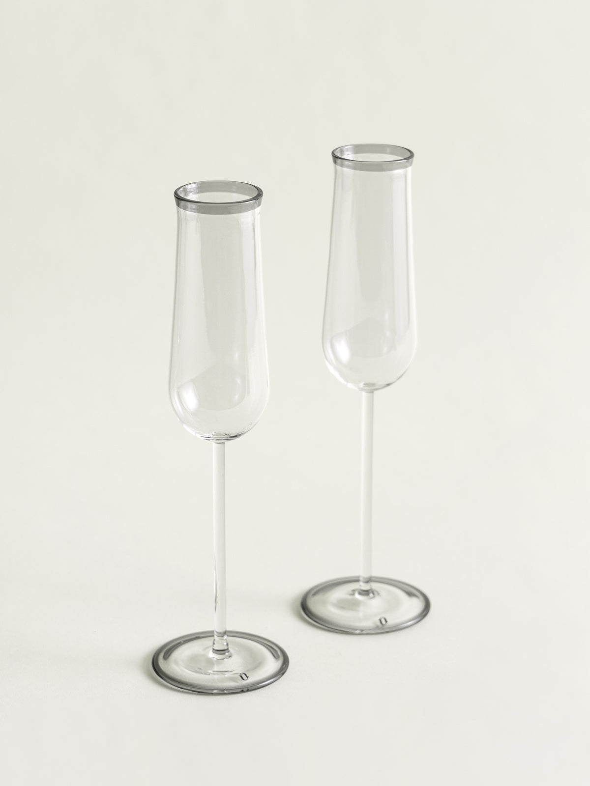 Two clear glass champagne flutes with a subtle gray rim, standing on a light cream background.
