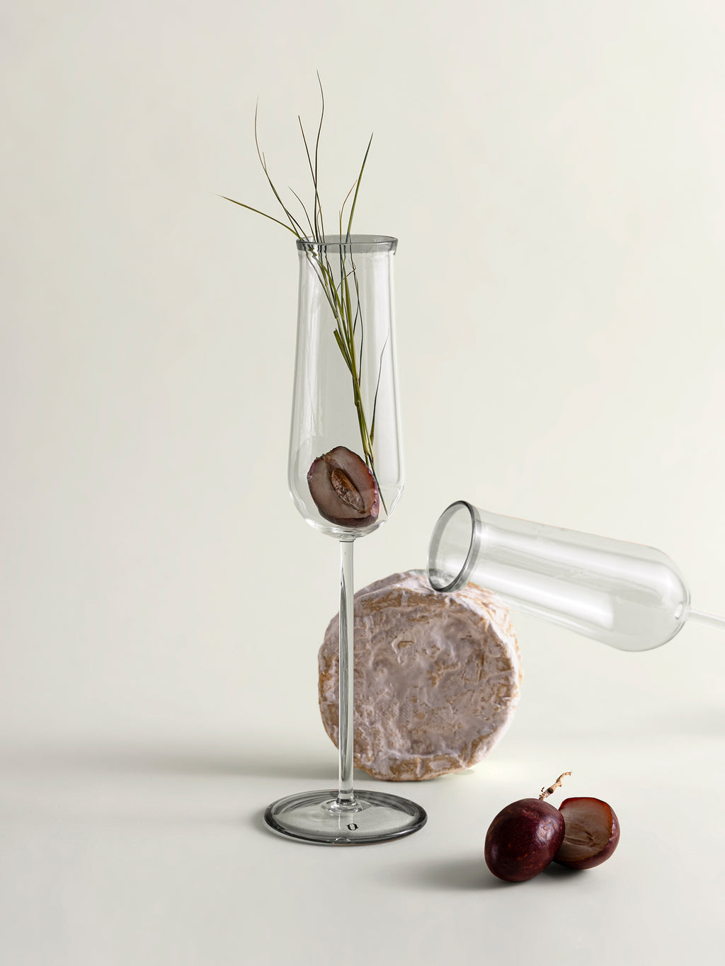 Two clear glass champagne flutes, styled with dates and a sprig of grass on a light beige background.