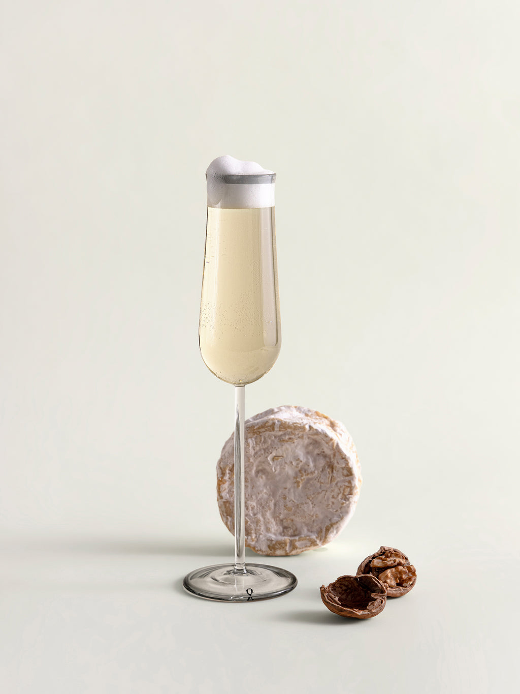 A clear glass flute with a grey rim, filled with a pale beverage, styled on a light neutral background with a cheese wheel and walnuts as props.