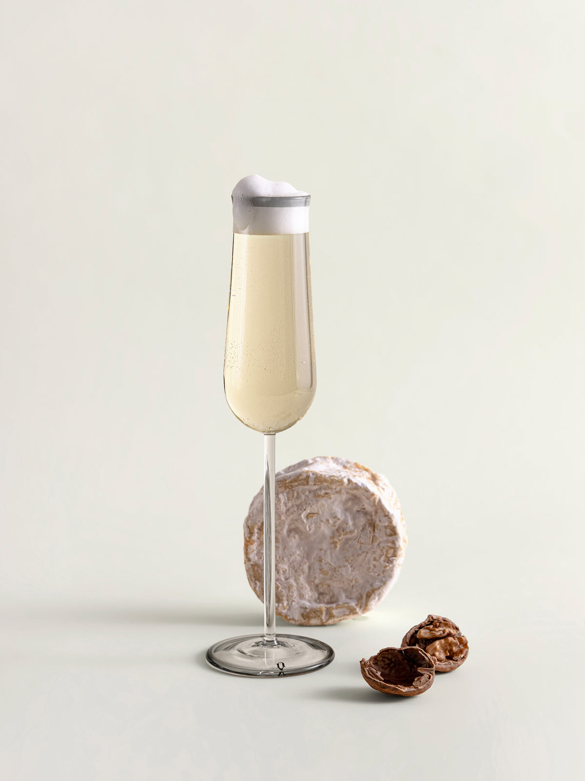 A clear glass flute with a grey rim, filled with a pale beverage, styled on a light neutral background with a cheese wheel and walnuts as props.