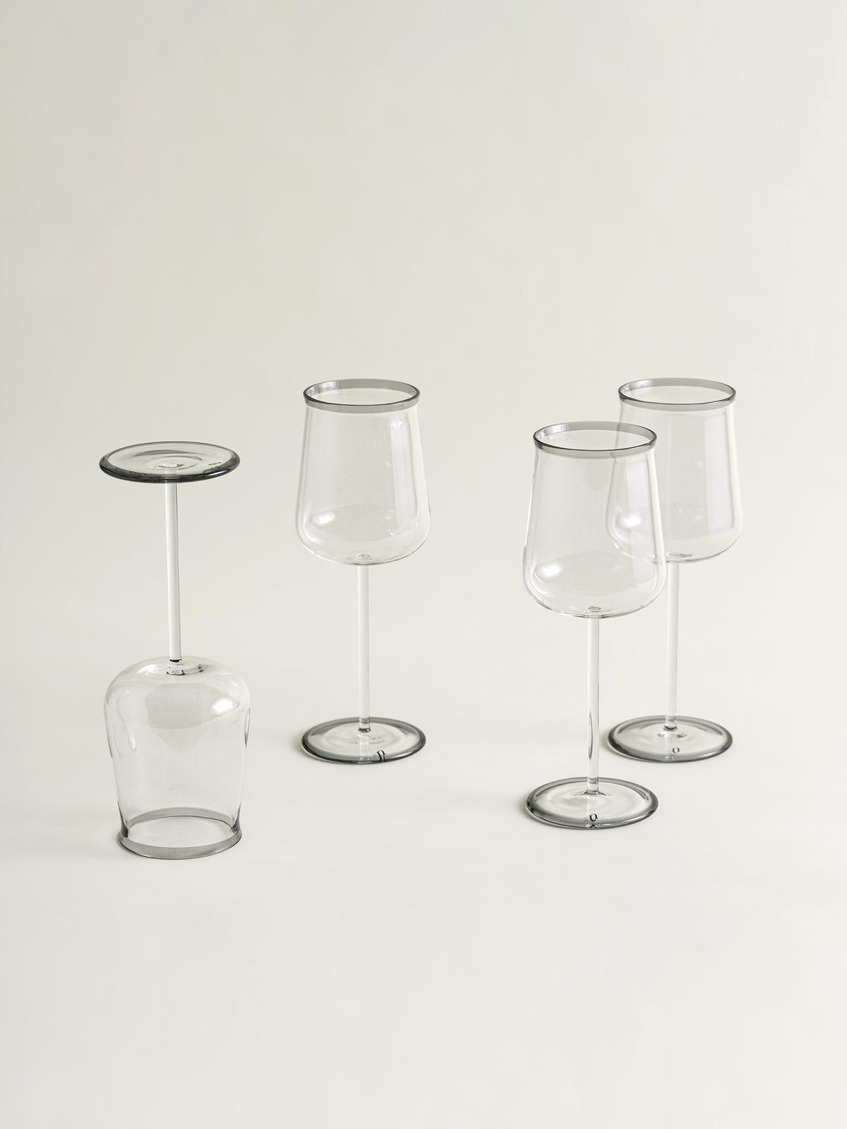 Four clear glass wine glasses with smoke grey rim accents arranged on a plain light background, including one tipped upside down.