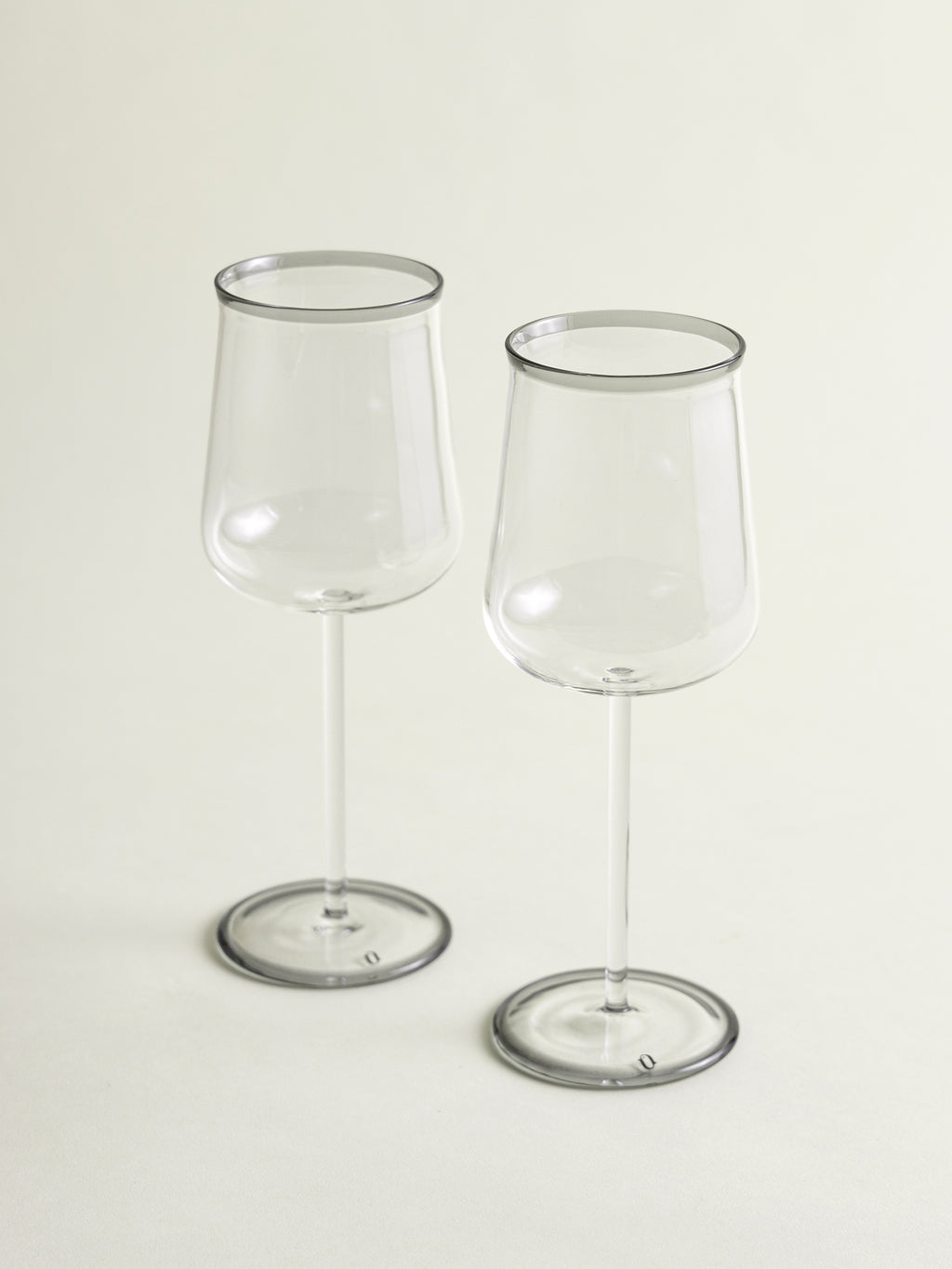 Two clear wine glasses with a smoke grey rim, standing on a light beige background.