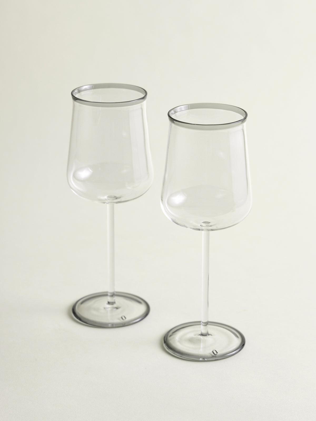 Two clear wine glasses with a smoke grey rim, standing on a light beige background.