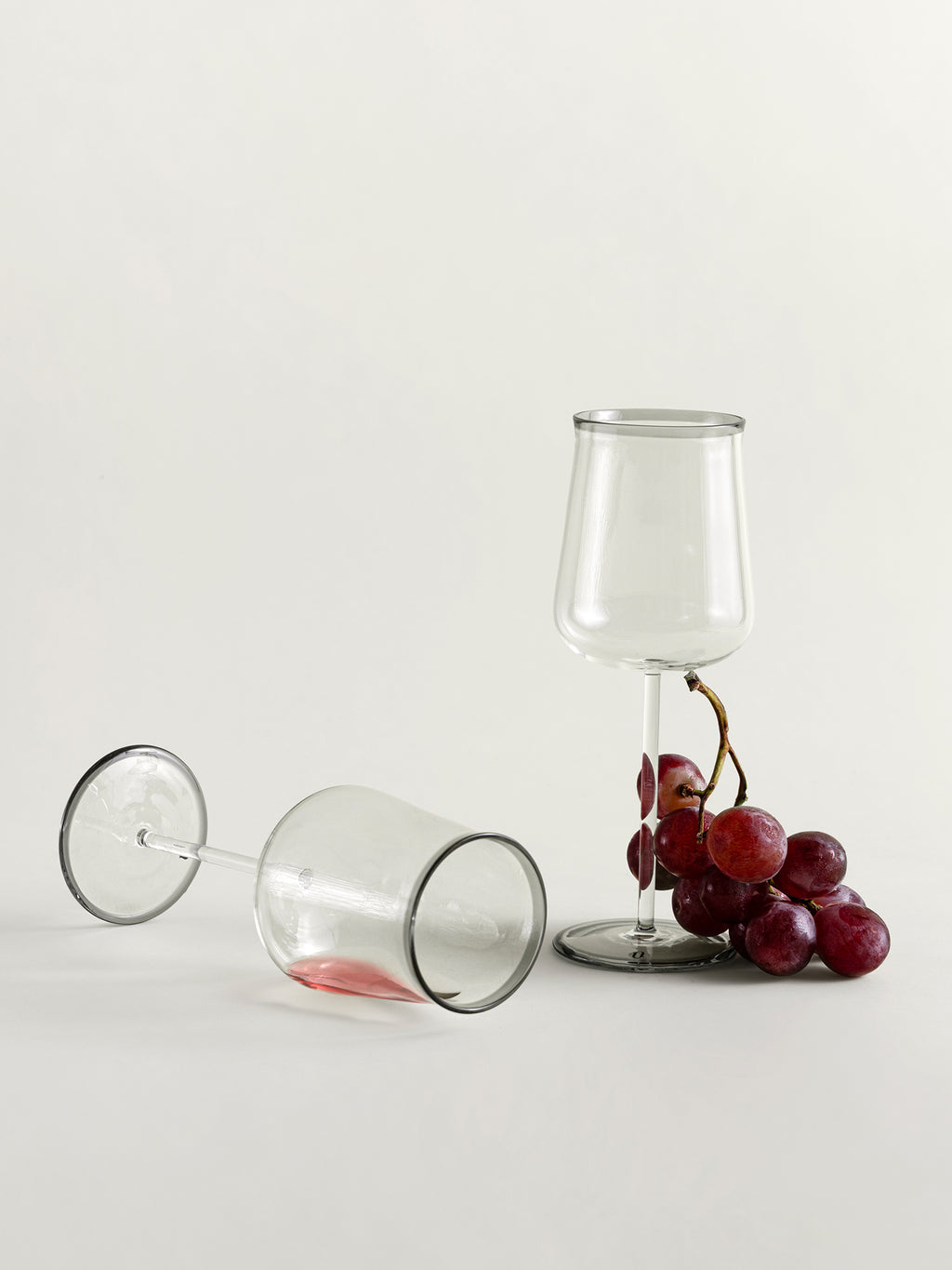 Two clear wine glasses on a light gray studio background, one standing upright and one lying on its side, with a cluster of red grapes beside the upright glass.