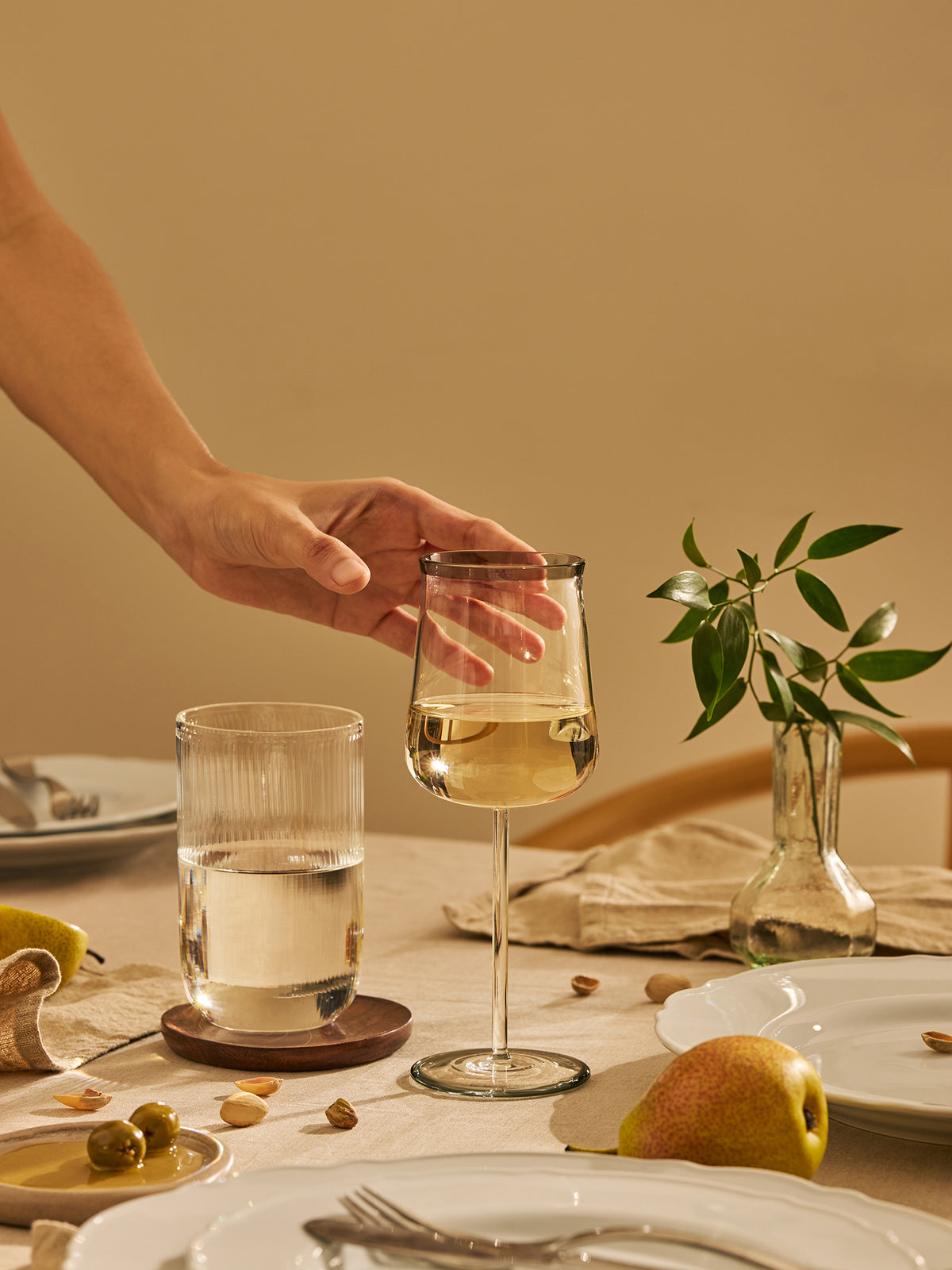 A hand approaching a clear wine glass with white wine in a warm, cozy dining room setting. The table is elegantly set with white plates, silver cutlery, a glass of water, a small vase with green leaves, and a pear.