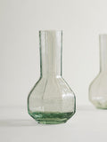 A clear textured glass vase with a bulbous base and narrow neck, shown on a light neutral background with a second blurred vase in the side.