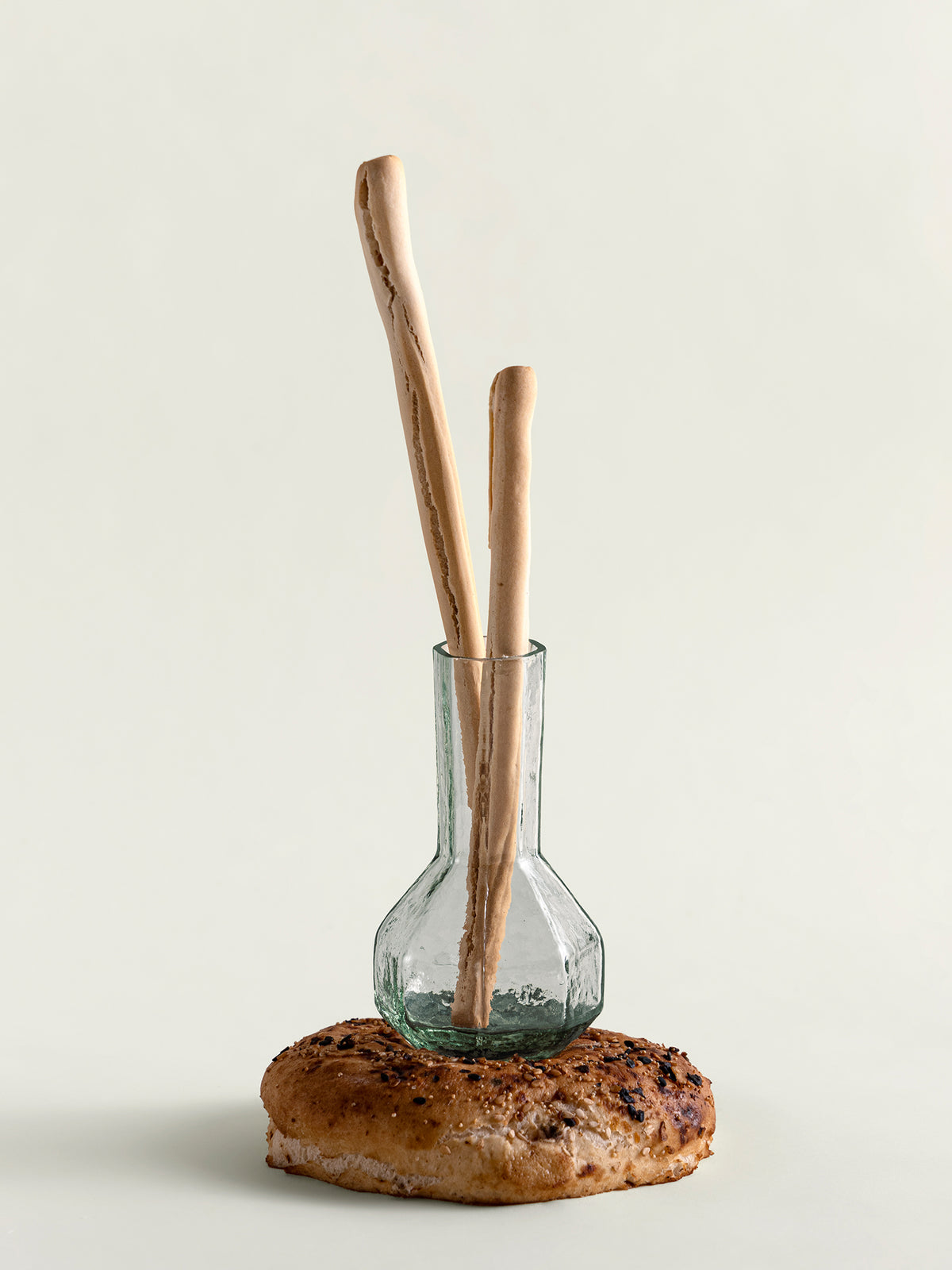 Two long wooden bread sticks standing upright in a small clear glass vase, which is placed on top of a round toasted bread slice against a plain light background.