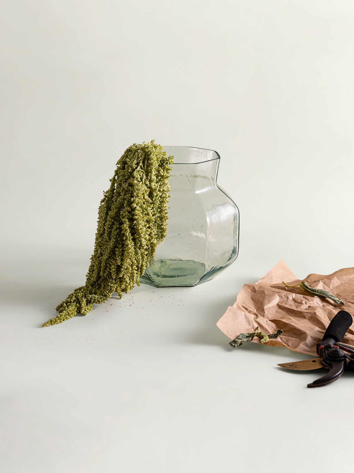 A clear glass vase with a wide flared rim and a narrow neck, styled on a light neutral background with dried green plant material draped over it and brown paper with scissors nearby.