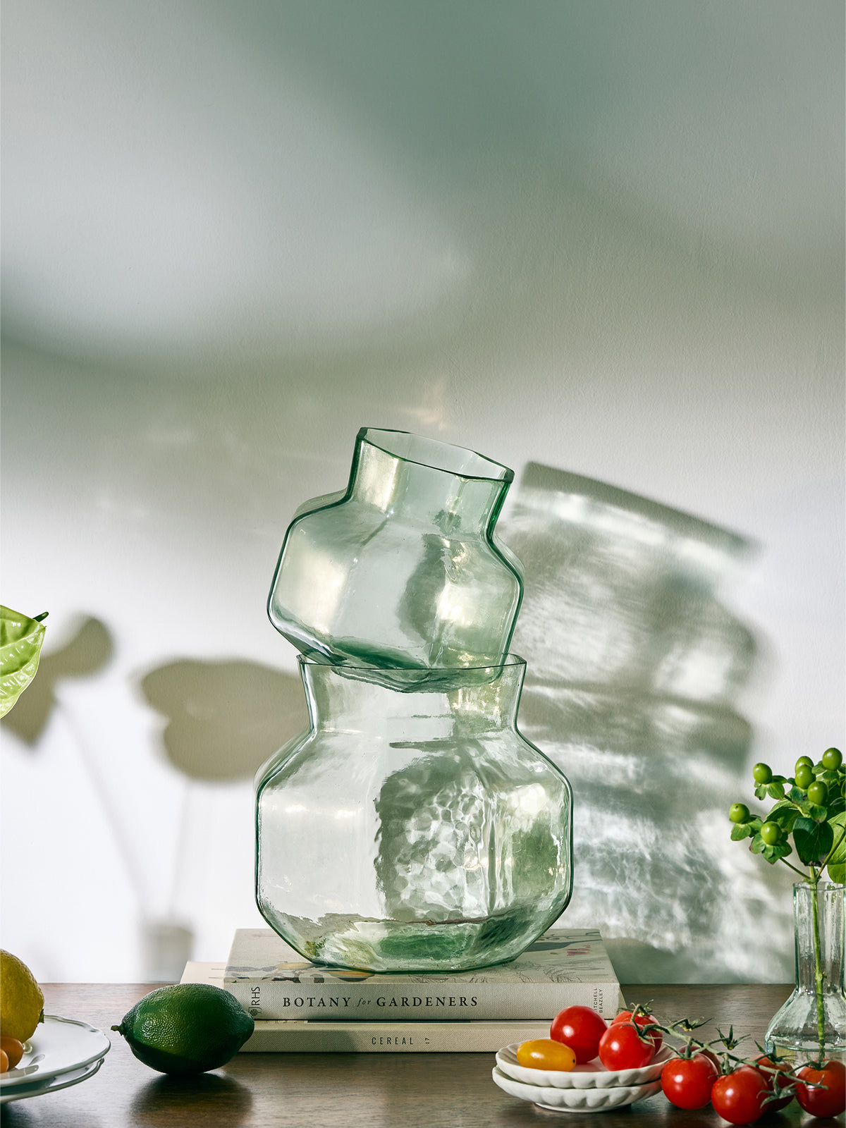 Clear glass vases with a bulbous base and a narrow neck, styled on a wooden table with books, tomatoes, and greenery in a softly lit interior setting.