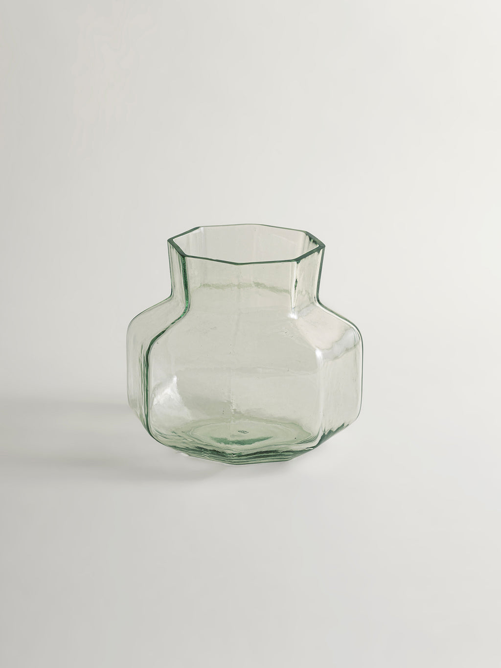 A small clear glass vase with a muted green tint, featuring a flared neck and a rounded, faceted base, set against a plain beige background.
