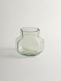 A small clear glass vase with a muted green tint, featuring a flared neck and a rounded, faceted base, set against a plain beige background.