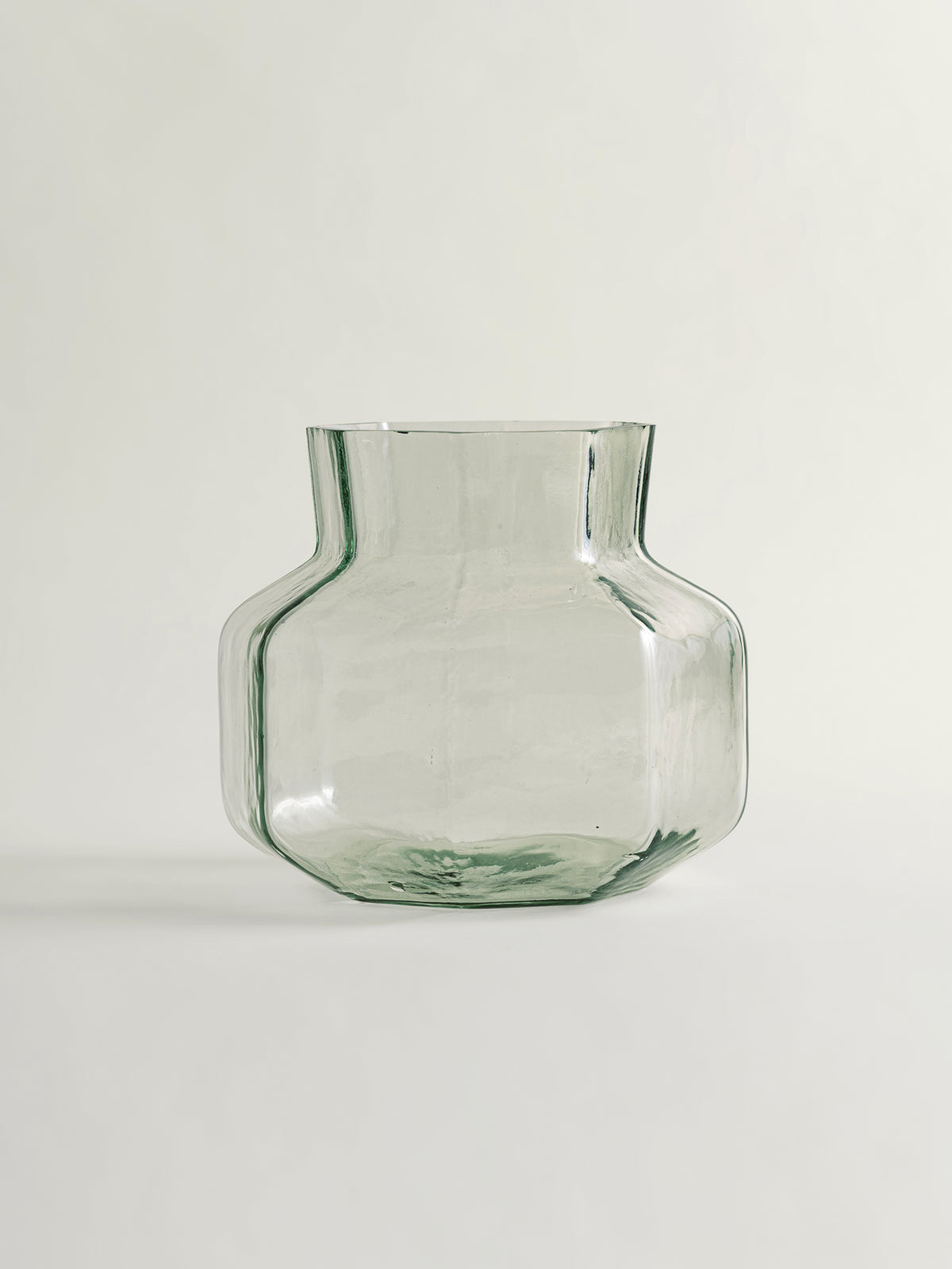 A clear light green glass vase with a wide, rounded body and a short, flared neck, photographed against a neutral off-white background.