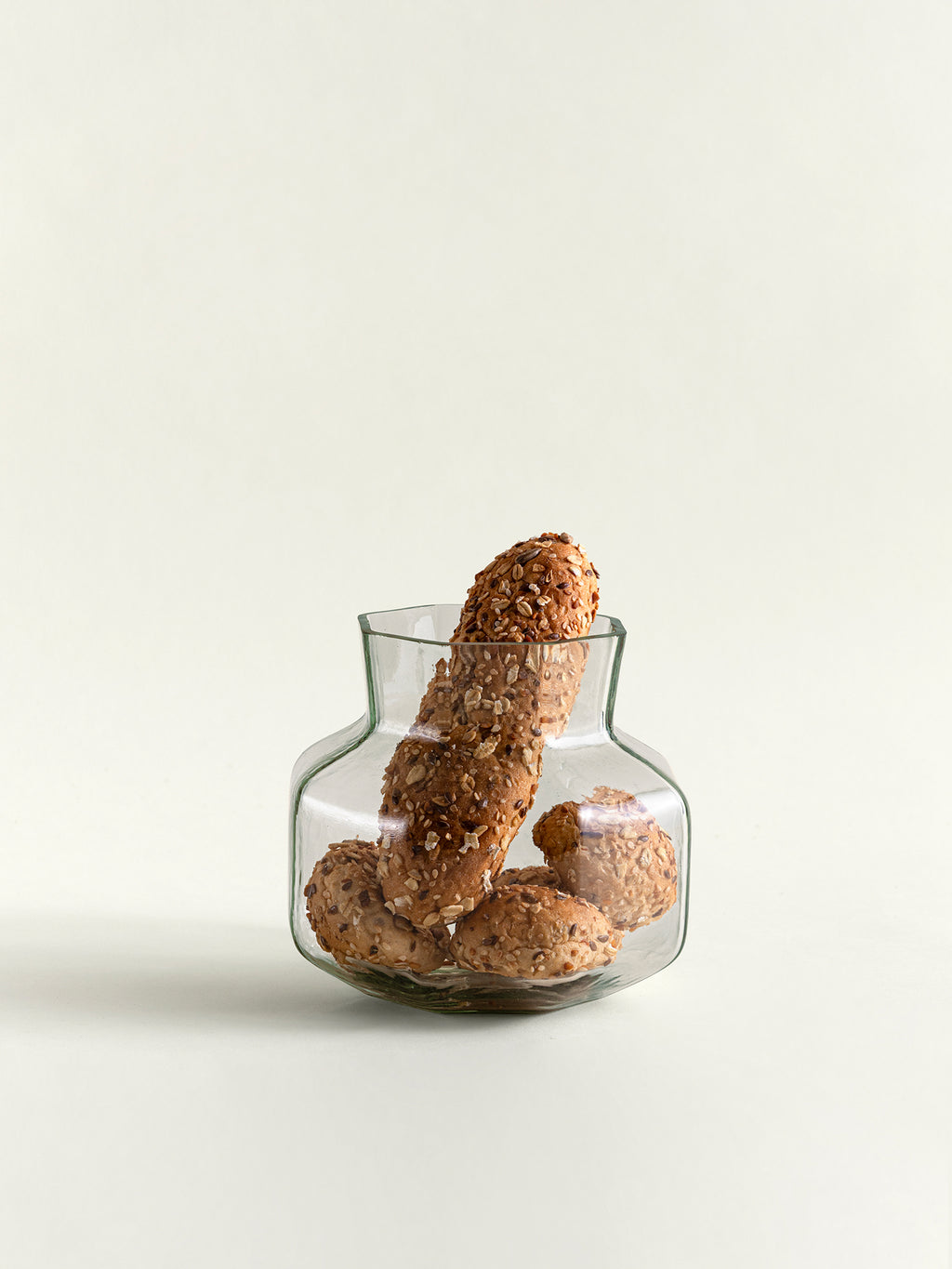 Several brown, sesame-seed-covered breads are arranged inside a small clear glass vase on a clean, light off-white background.
