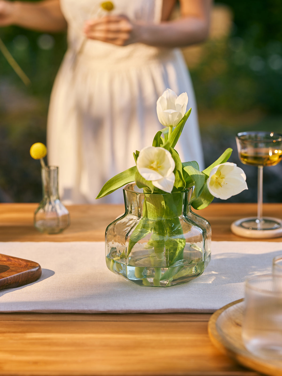 A woman in a white sleeveless dress stands blurred in the background holding small yellow flowers, while a glass vase with white tulips sits on a wooden table in the foreground. The scene is set outdoors on a wooden dining table with a white runner, a wine glass, and plates.