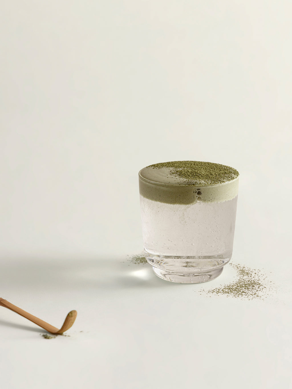 A clear glass filled with soda, topped with a matcha foam and dusted matcha, set on a light neutral background with scattered powder around the base and a chasen visible in the lower left.
