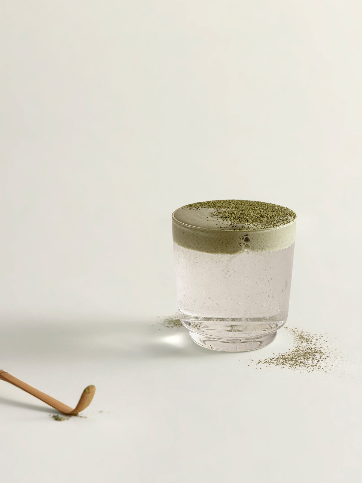 A clear glass filled with soda, topped with a matcha foam and dusted matcha, set on a light neutral background with scattered powder around the base and a chasen visible in the lower left.