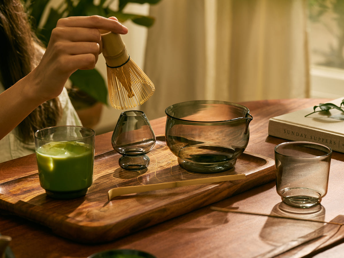A person's hand holds a bamboo whisk above a glass of matcha on a wooden tray, with matching glassware and a bamboo scoop arranged on a wooden table in a warm, softly lit interior setting.