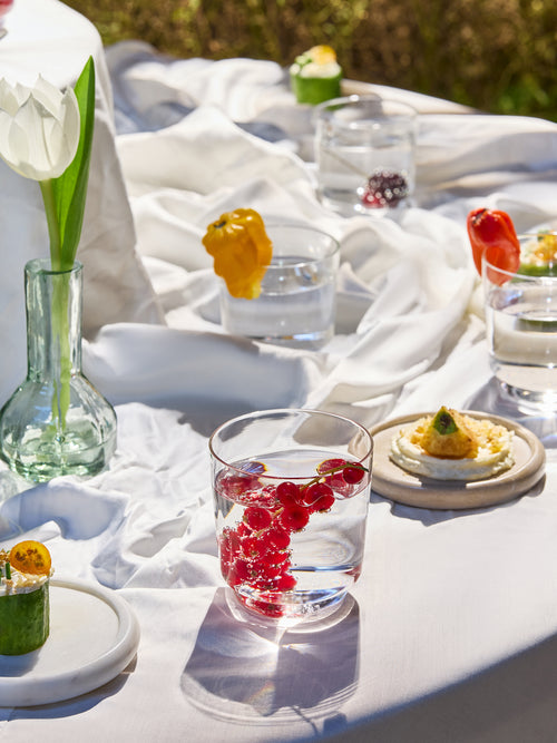 A styled outdoor table setting with a white tablecloth, clear glass cups containing red berries, small plates with appetizers, a green glass vase with a white tulip, and blurred greenery in the background.