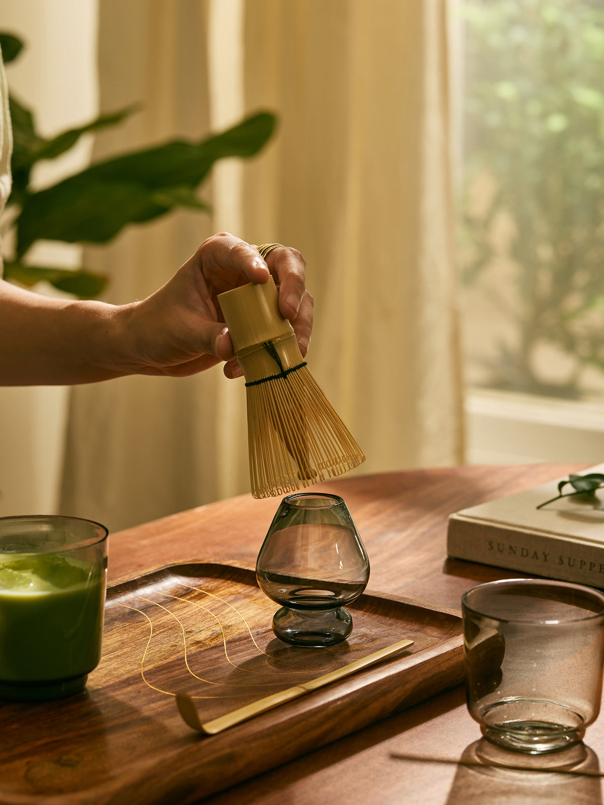 A hand holds a bamboo whisk above a glass matcha whisk stand on a wooden tray, with a green drink in a glass and an empty glass cup nearby on a warm indoor table setting.