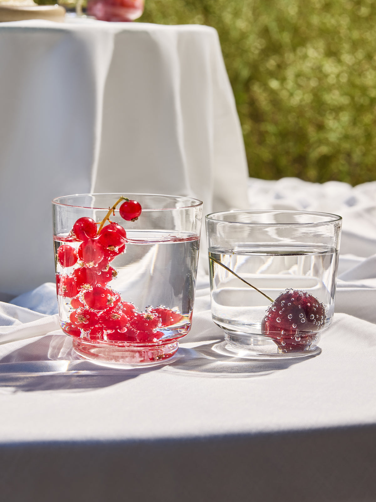 Two clear glass tumblers filled with water on a white tablecloth outdoors, one containing red berries and the other a dark berry, with a blurred green garden background.