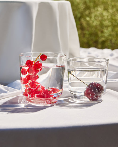 Two clear glass tumblers filled with water on a white tablecloth outdoors, one containing red berries and the other a dark berry, with a blurred green garden background.