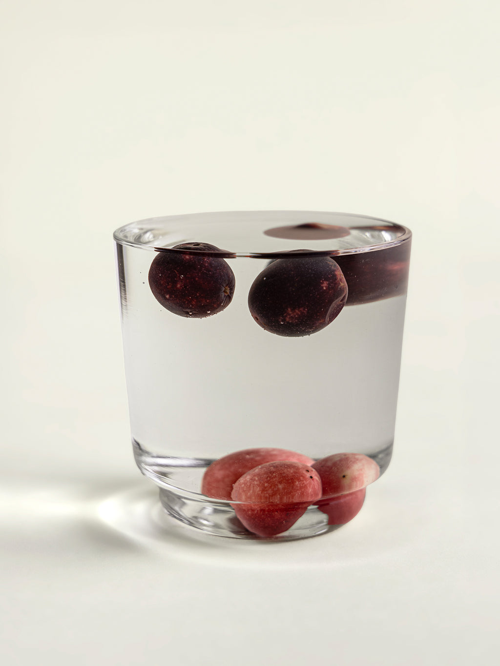 A clear glass tumbler filled with clear water, containing dark red karondas floating near the top and several others settled at the bottom, photographed on a clean white background.