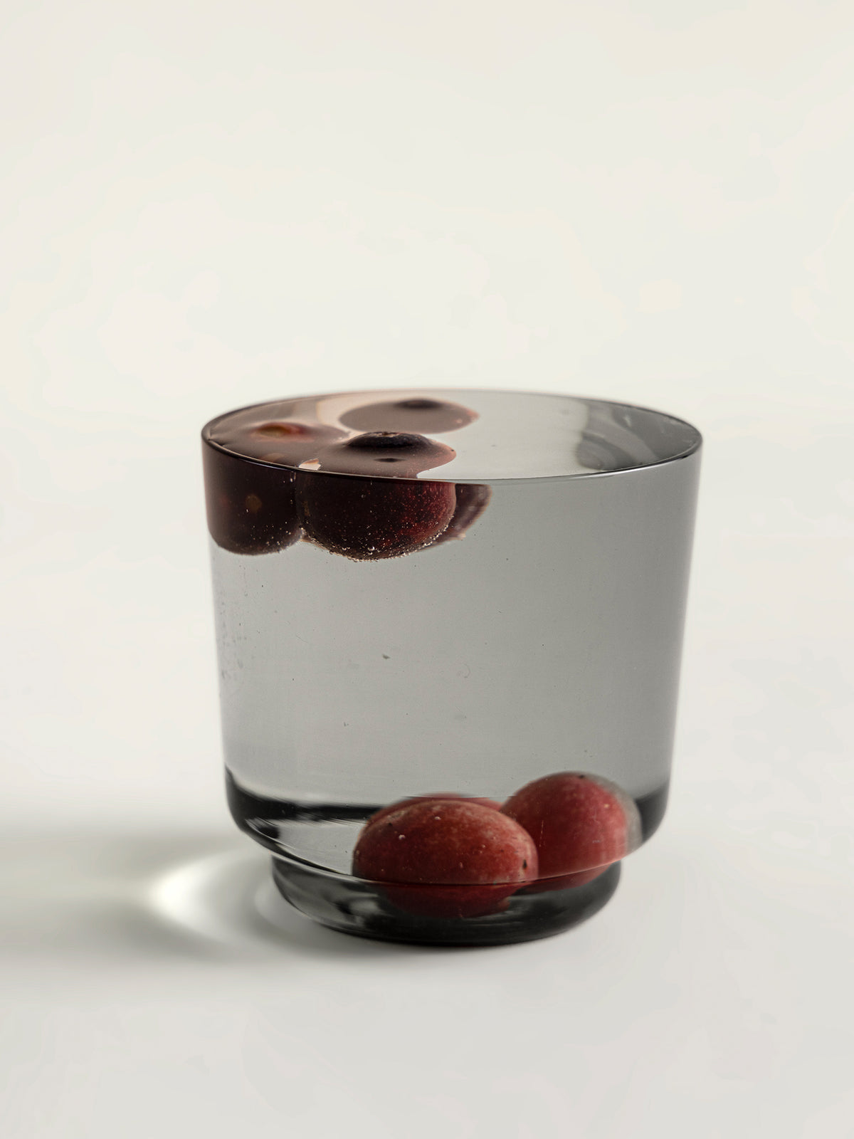 A smoke grey glass tumbler with a thick base, containing water and several red karondas, photographed on a clean white background.