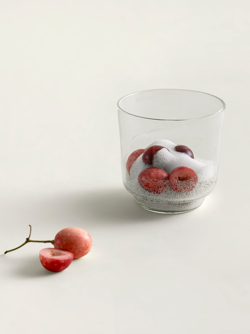 A clear glass containing red karondas and white foam, photographed on a clean off-white surface with a couple of loose karondas beside it.