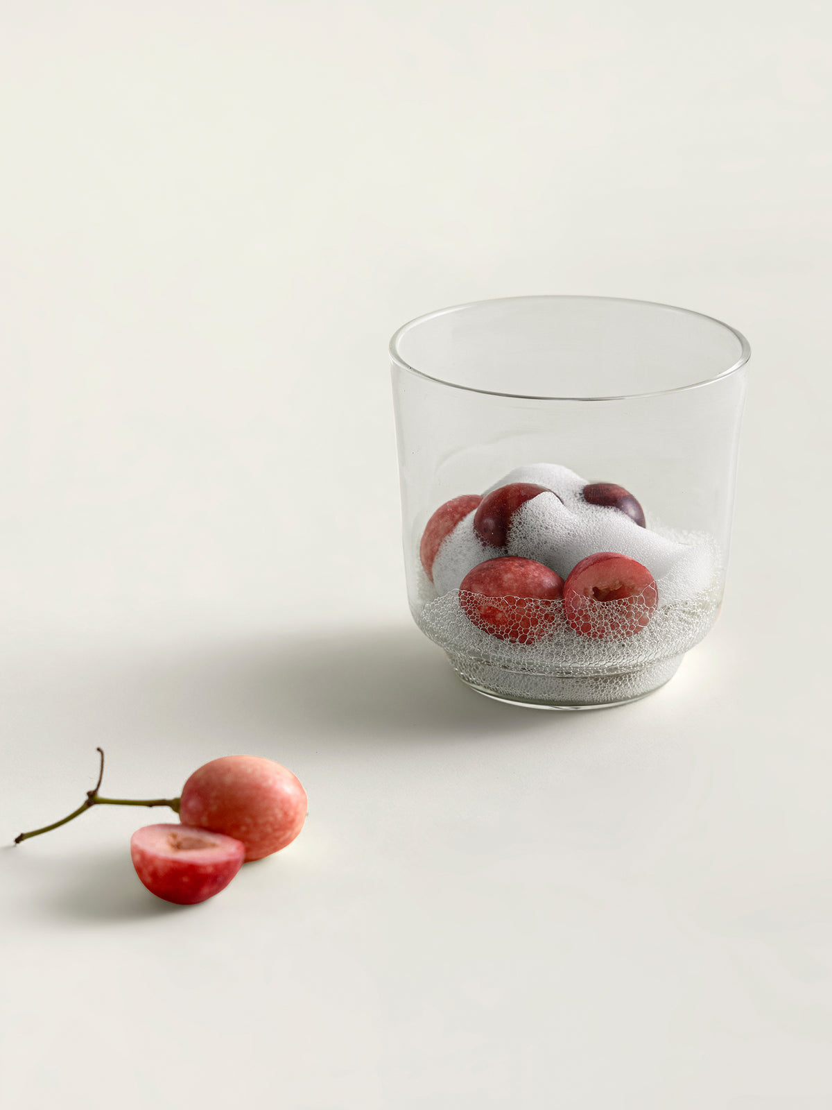A clear glass containing red karondas and white foam, photographed on a clean off-white surface with a couple of loose karondas beside it.
