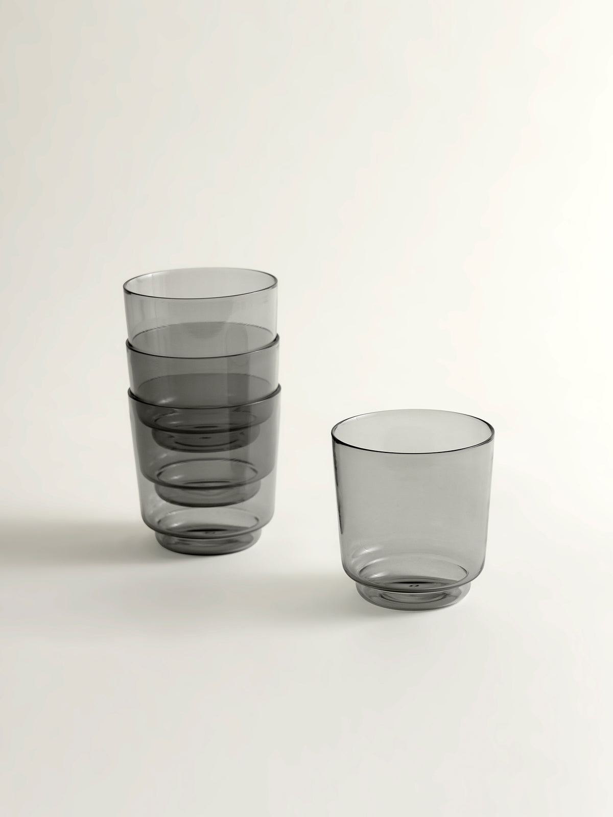Four translucent smoke grey glass tumblers with a modern tapered shape, shown on a clean off-white background.
