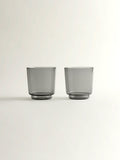 Two translucent smoke grey glass tumblers with a modern tapered shape, shown on a clean off-white background.