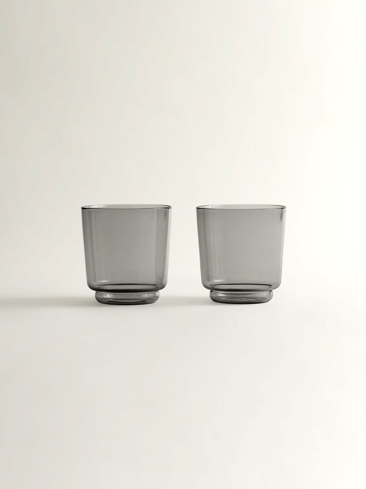 Two translucent smoke grey glass tumblers with a modern tapered shape, shown on a clean off-white background.