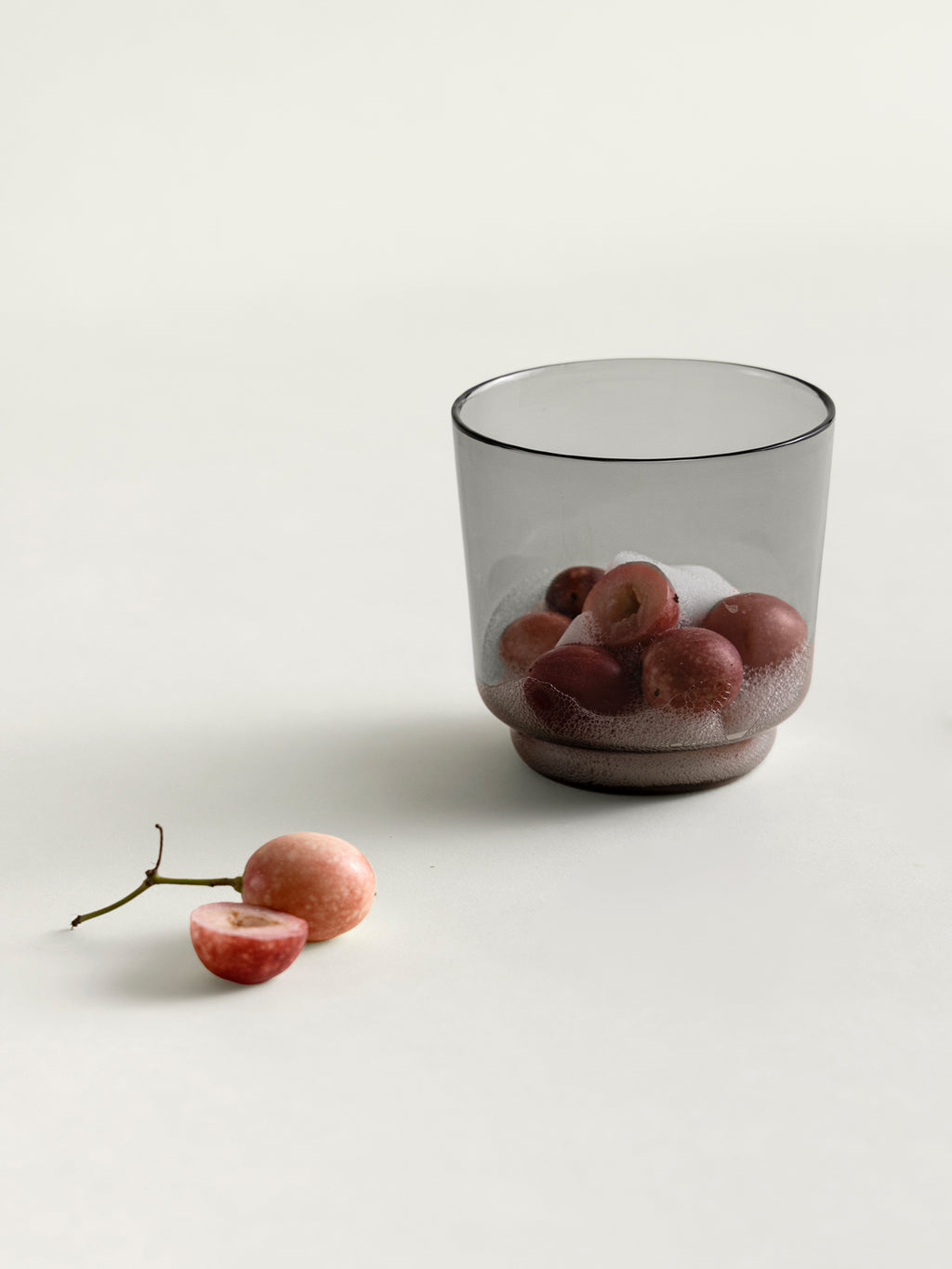 A translucent smoke grey tumbler glass containing karonda, with a few karondas placed beside it on a clean off-white surface.