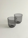 Two translucent smoke grey glass tumblers with a modern tapered shape, shown on a clean off-white background.