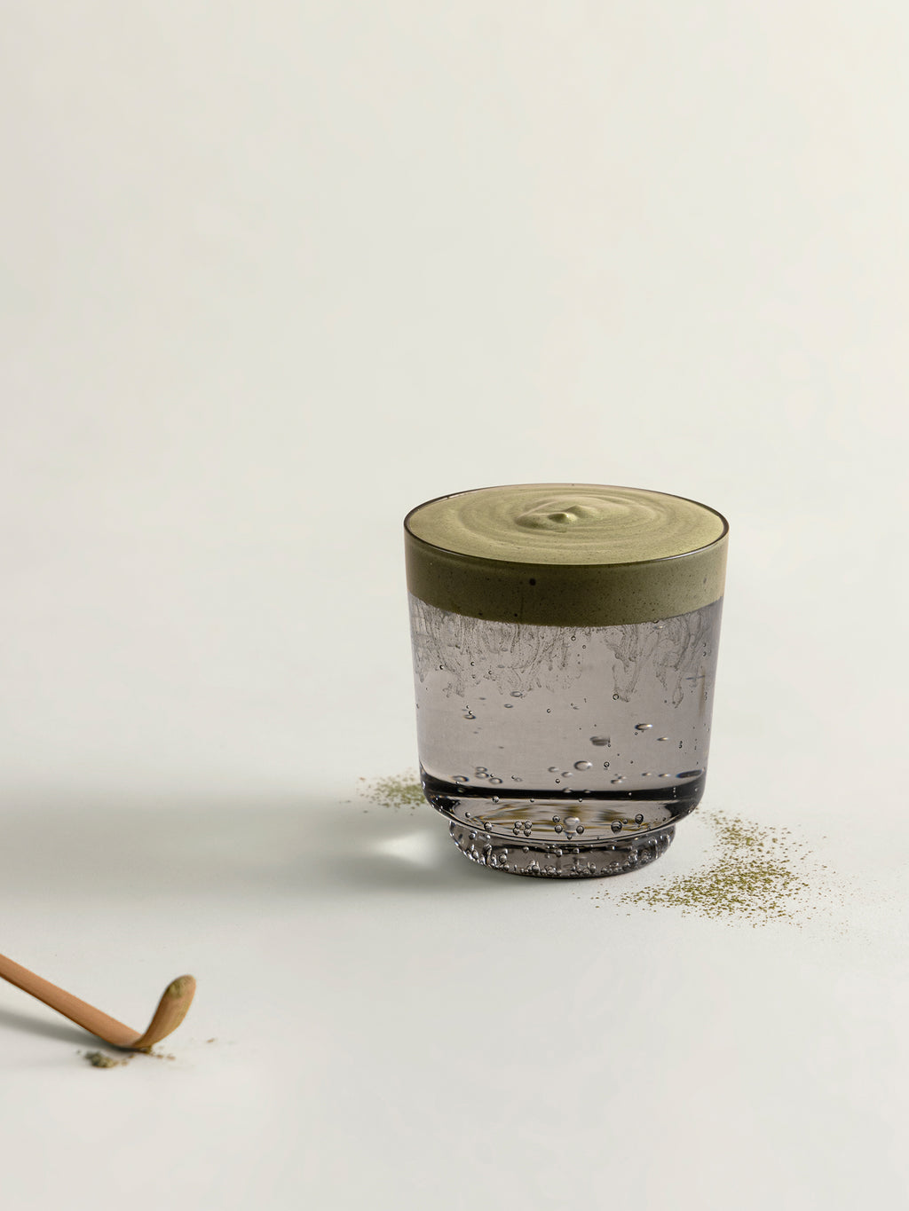 A clear glass tumbler with sparkling water and matcha foam, styled on a light beige surface with scattered dried matcha and a wooden spoon handle visible in the corner.