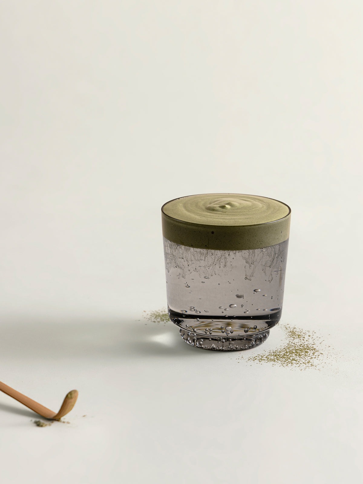 A clear glass tumbler with sparkling water and matcha foam, styled on a light beige surface with scattered dried matcha and a wooden spoon handle visible in the corner.