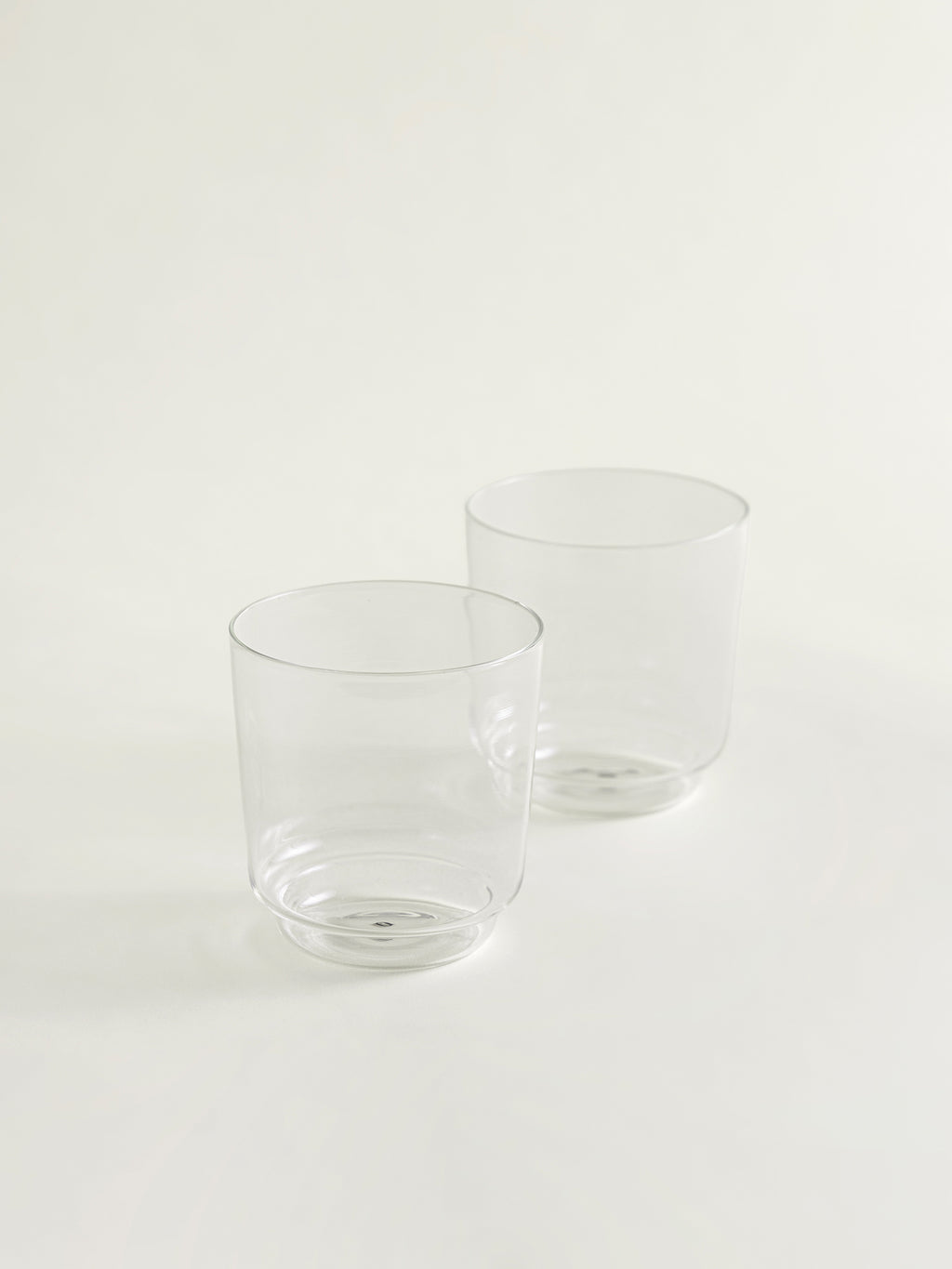 Two clear glass tumblers with thick and round bases, arranged on a seamless off-white background.