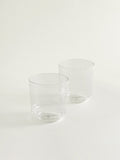 Two clear glass tumblers with thick and round bases, arranged on a seamless off-white background.