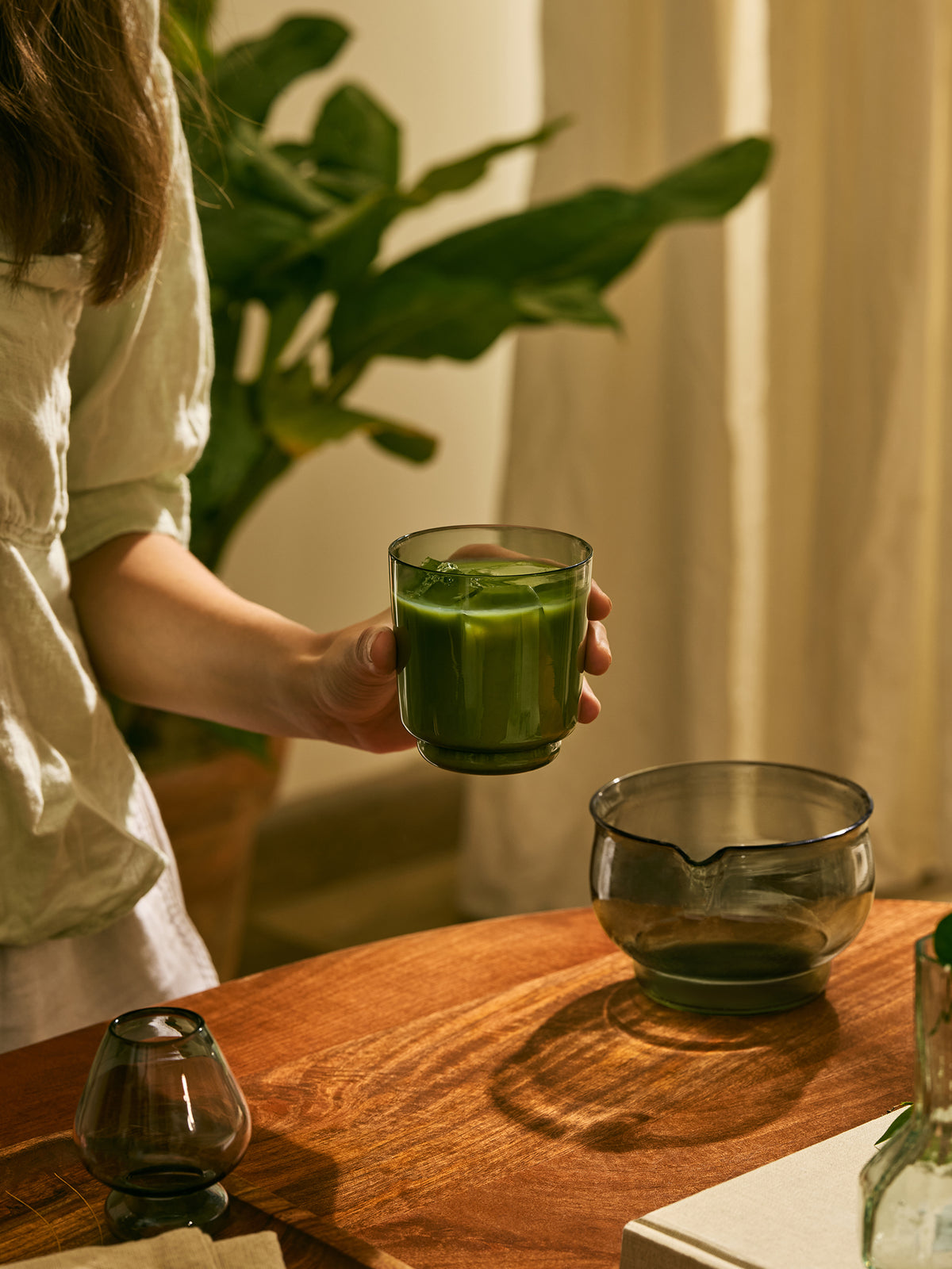 A person in a white shirt holds a grey glass tumbler filled with green matcha tea over a wooden table, with a matching glass bowl and matcha whisk stand nearby in a warm indoor setting.