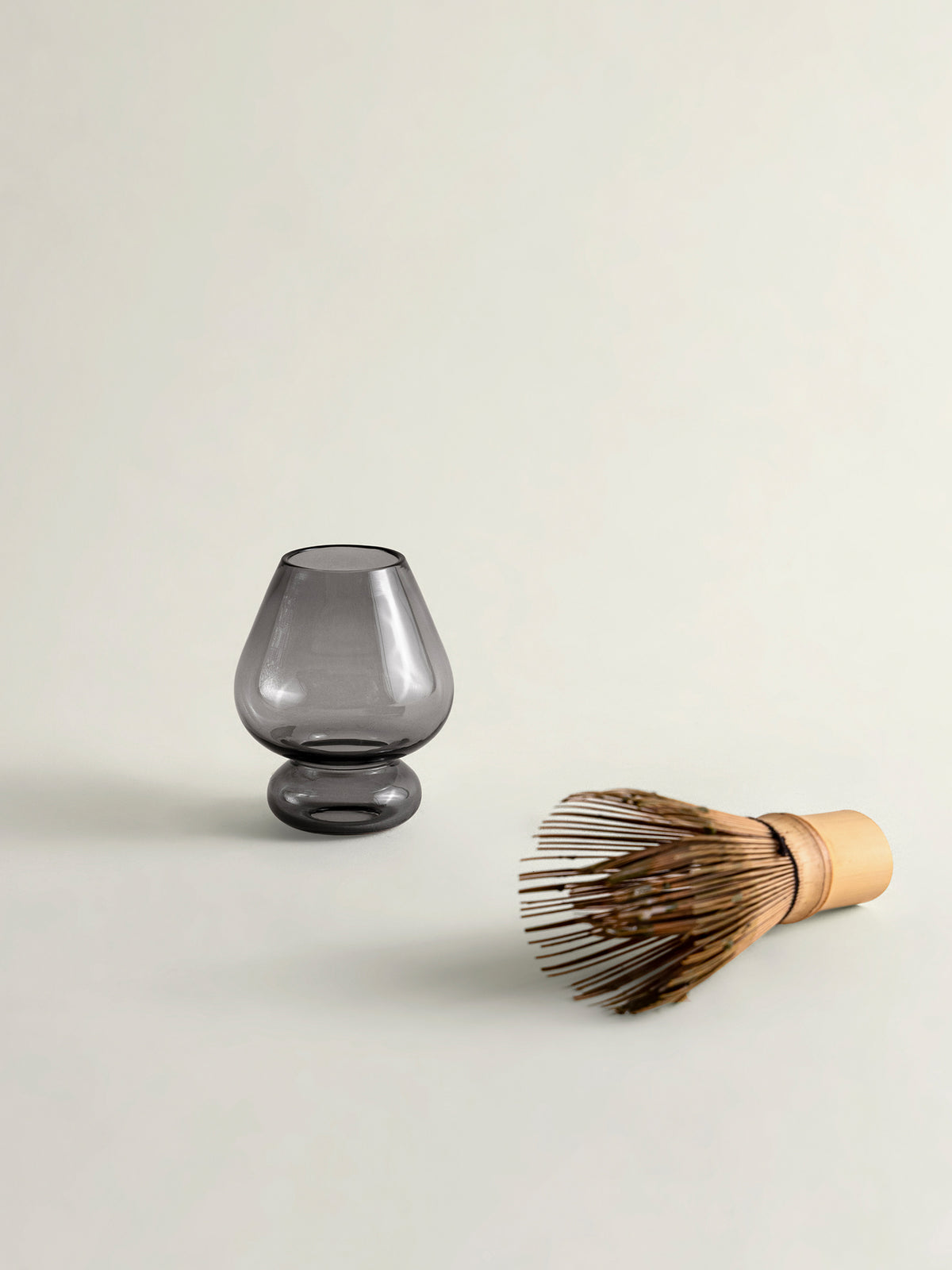 A smoke grey translucent glass matcha whisk holder with a bulbous shape and a bamboo whisk with brown bristles and a light wood handle, arranged on a clean off-white background.