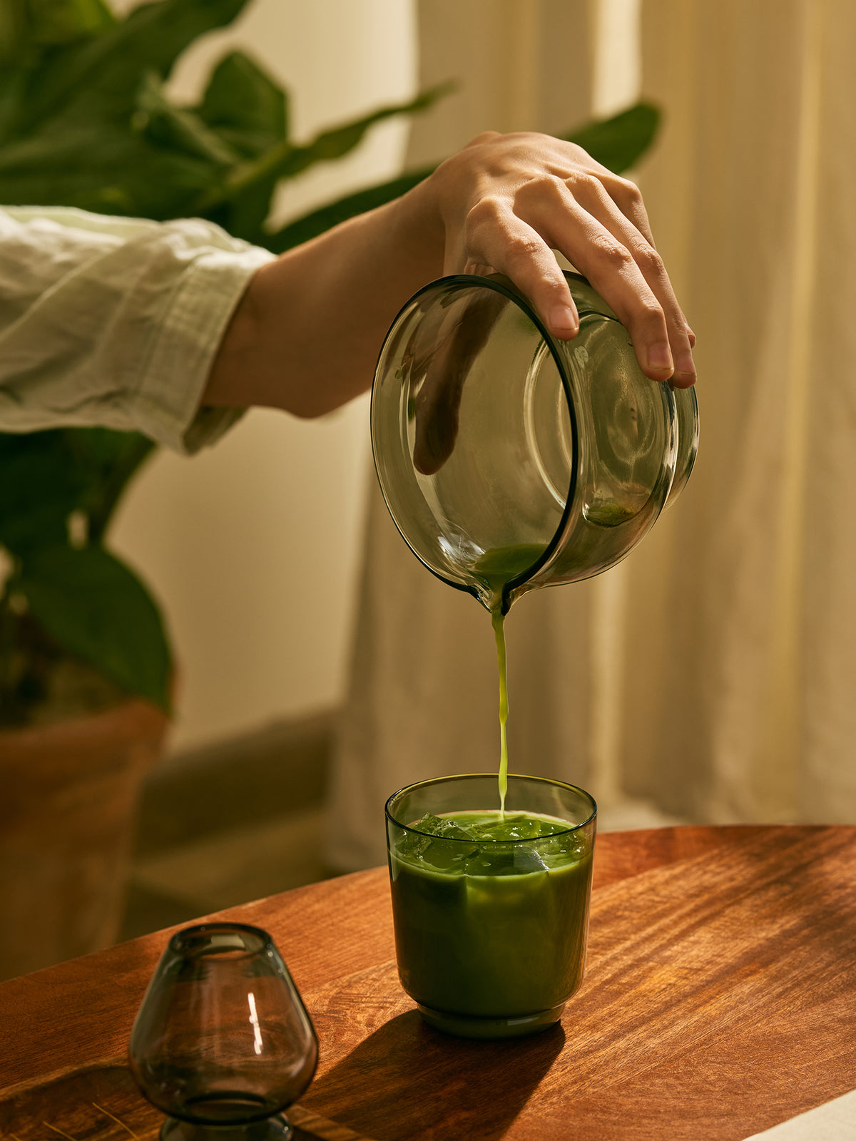 A hand pours green matcha tea from a clear glass pitcher into a glass cup on a wooden table, with a potted plant and curtains in the background.