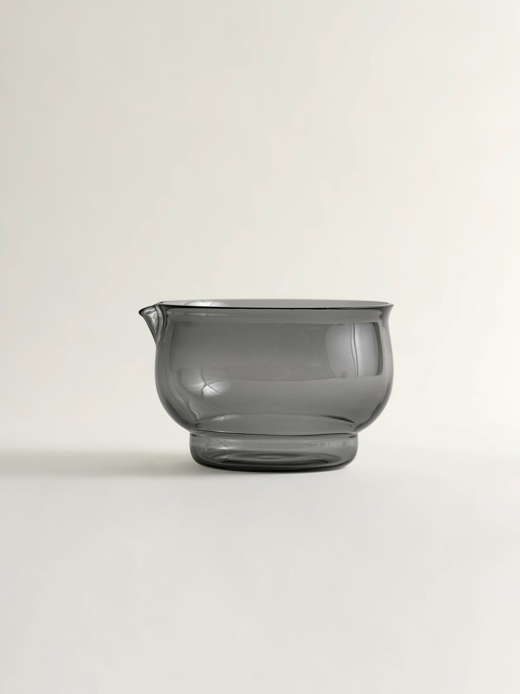 a smoke grey translucent glass bowl with a small pouring spout, photographed on a clean off-white background.
