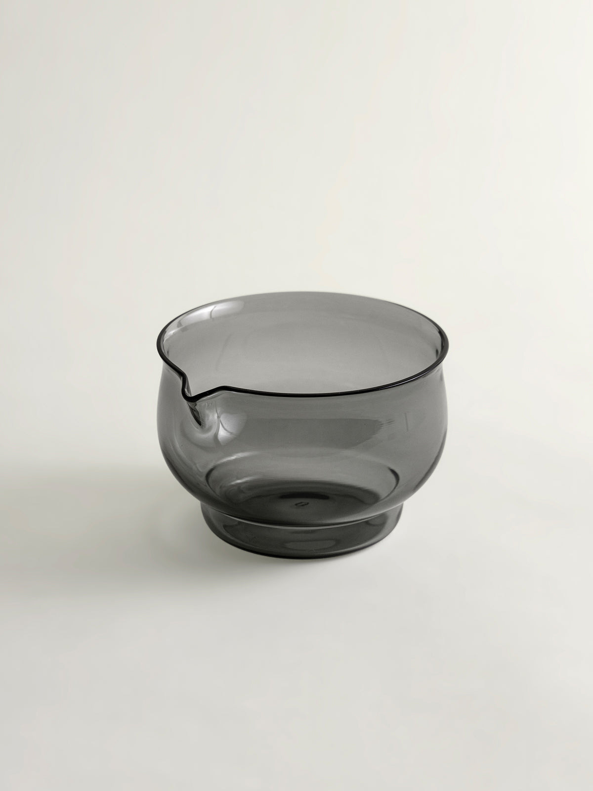 A smoke grey translucent glass bowl with a small pouring spout, shown on a plain light background.