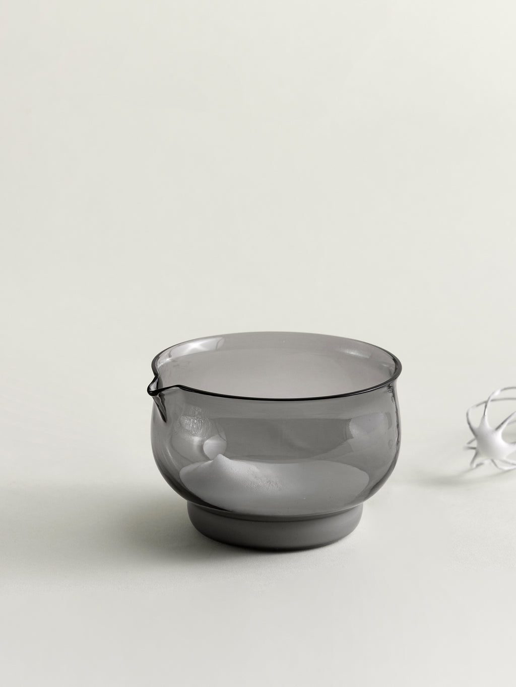 A translucent smoke grey glass bowl with a small spout on the left side, shown on a clean off-white background with a small white decorative object partially visible on the right.