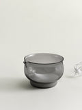 A translucent smoke grey glass bowl with a small spout on the left side, shown on a clean off-white background with a small white decorative object partially visible on the right.