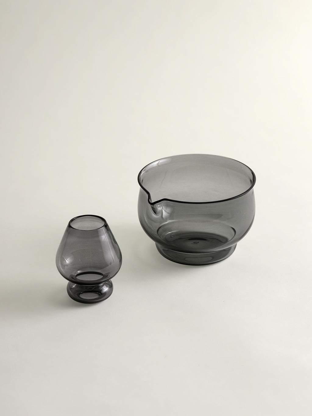 Two translucent smoky gray glass vessels on a clean off-white background, including a small whisk holder and a larger whisking bowl with a small pouring spout.
